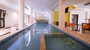 Zona de piscina interior moderna con una piscina larga y estrecha, una zona de estar con sillas naranjas y vistas a un jardín a través de un muro de cristal.