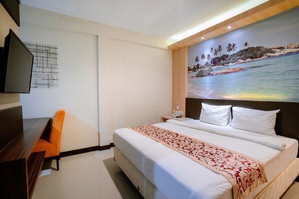 A hotel room with a bed, a desk, an orange chair, a painting of a beach scene, and a TV mounted on the wall.