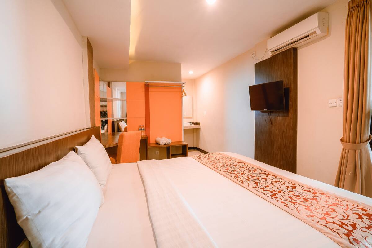 A hotel room with a bed, a television, an air conditioning unit, and a bathroom visible in the background.