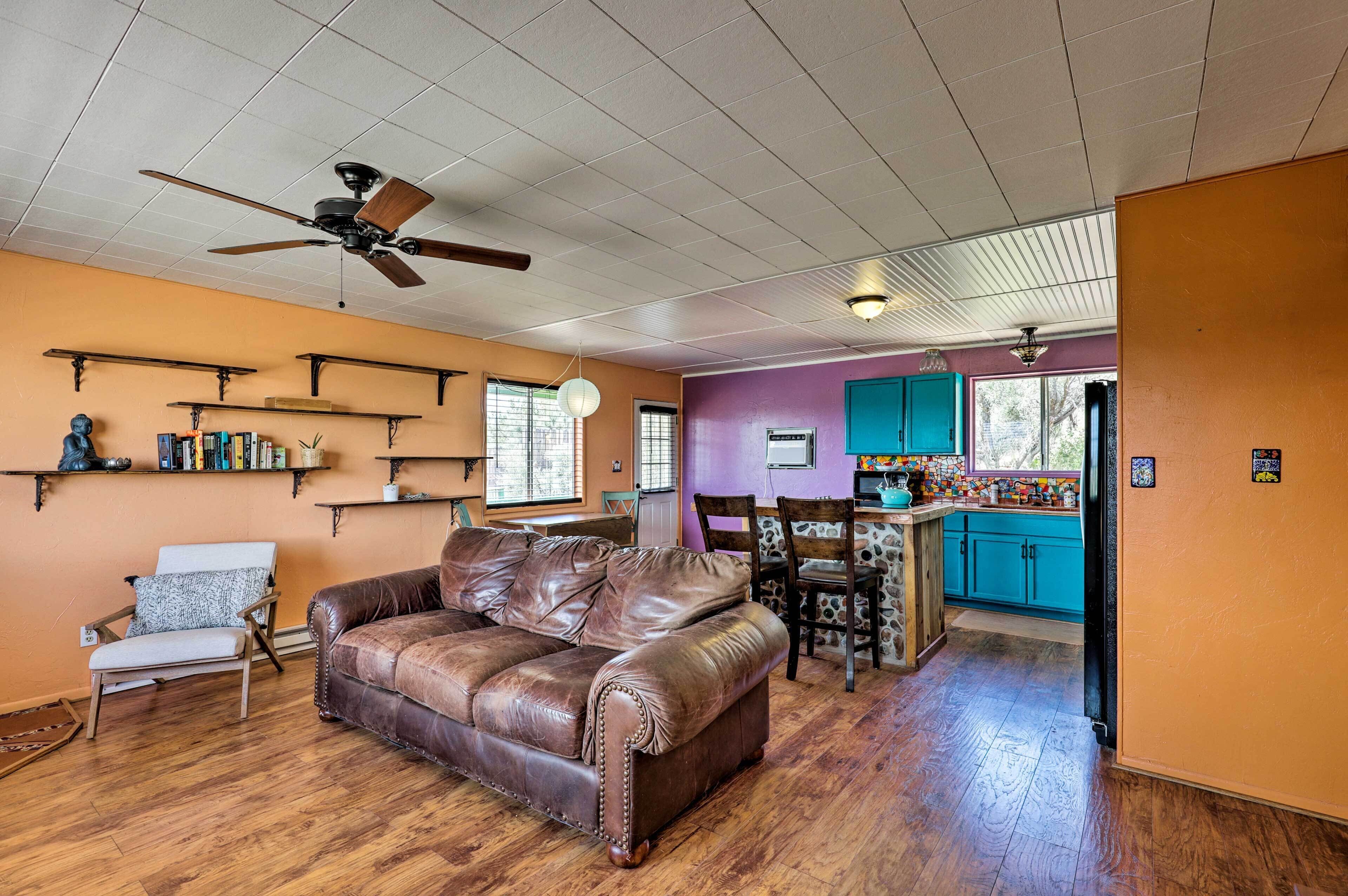 A living room with a brown leather couch, wooden floor, ceiling fan, and a kitchen area with teal cabinets and a window.
