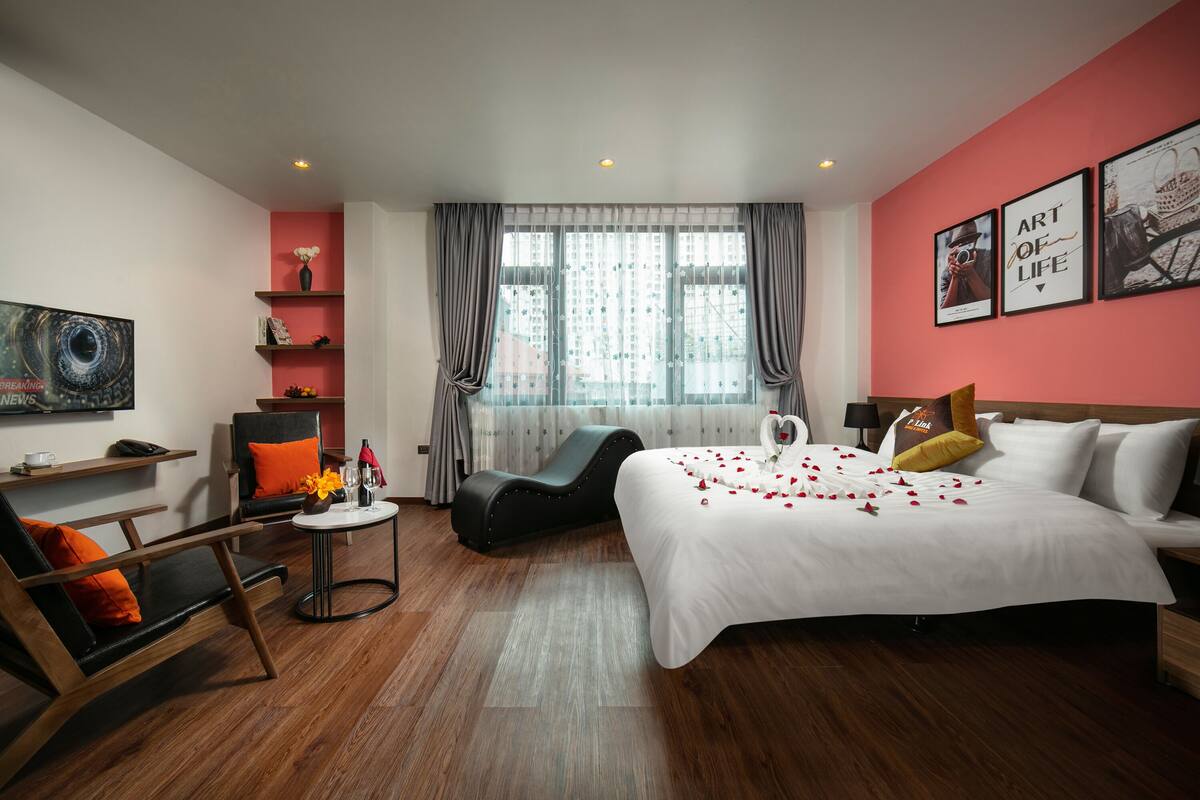 A modern hotel room with a large bed, a sofa, a small table, and a chair. There is a window with curtains, a wall with framed pictures, and a shelf with decorative items.
