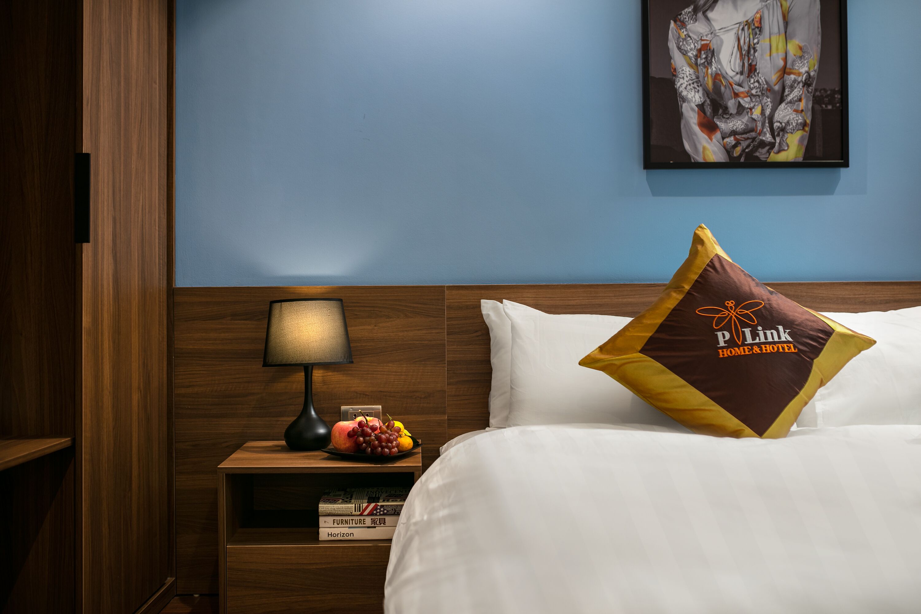A hotel room with a wooden headboard, a bedside table with a lamp, a fruit bowl, and a pillow with a logo.