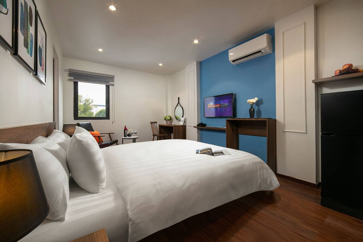 A hotel room with a bed, a desk, a chair, a TV, and an air conditioning unit.