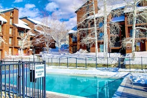 Zona de piscina con un cartel de advertencia para socorristas, rodeada de edificios cubiertos de nieve y árboles sin hojas.