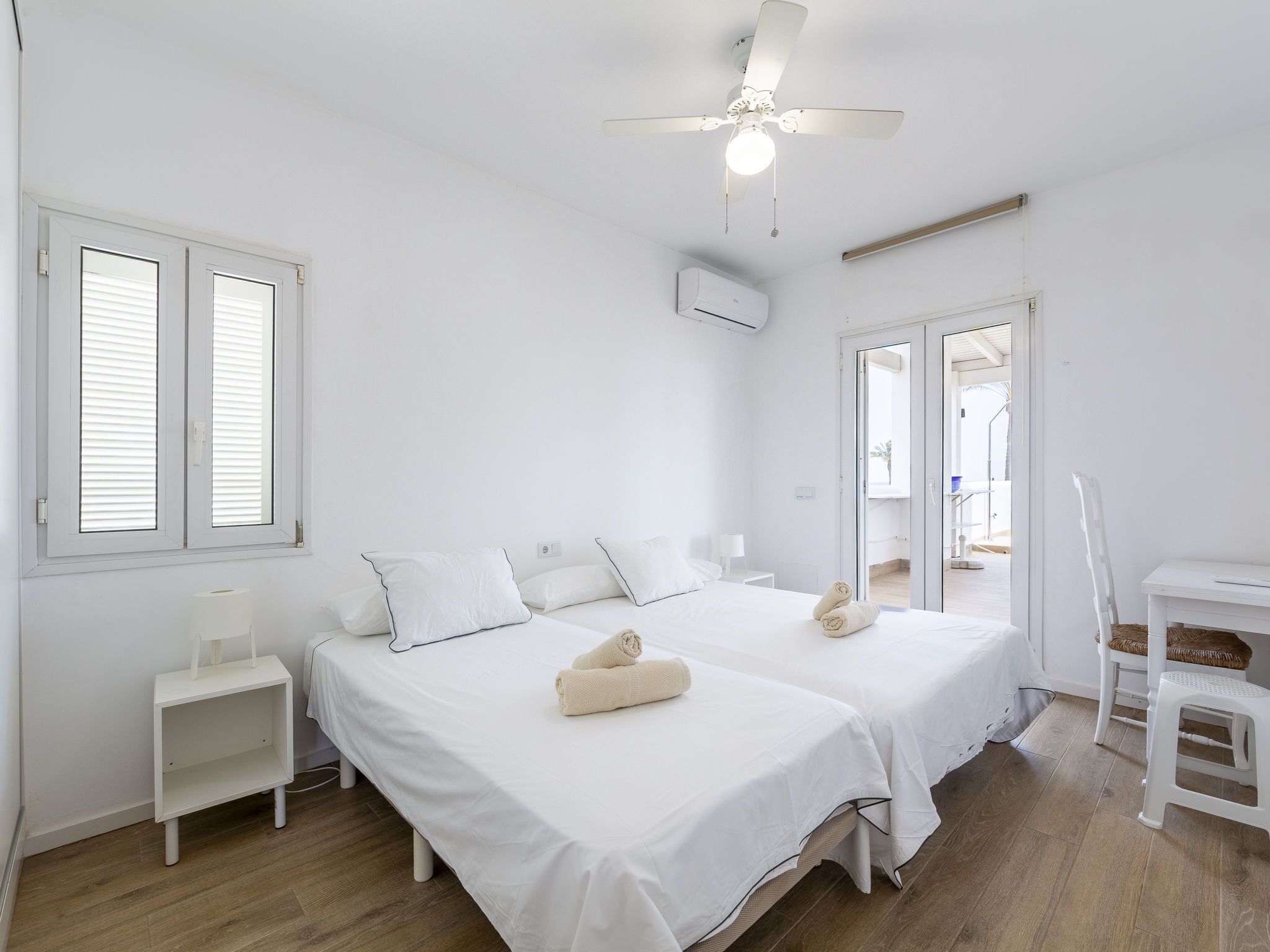 A clean, white room with two beds, a ceiling fan, and a view to the outside.