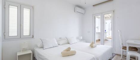 A clean, white room with two beds, a ceiling fan, and a view to the outside.