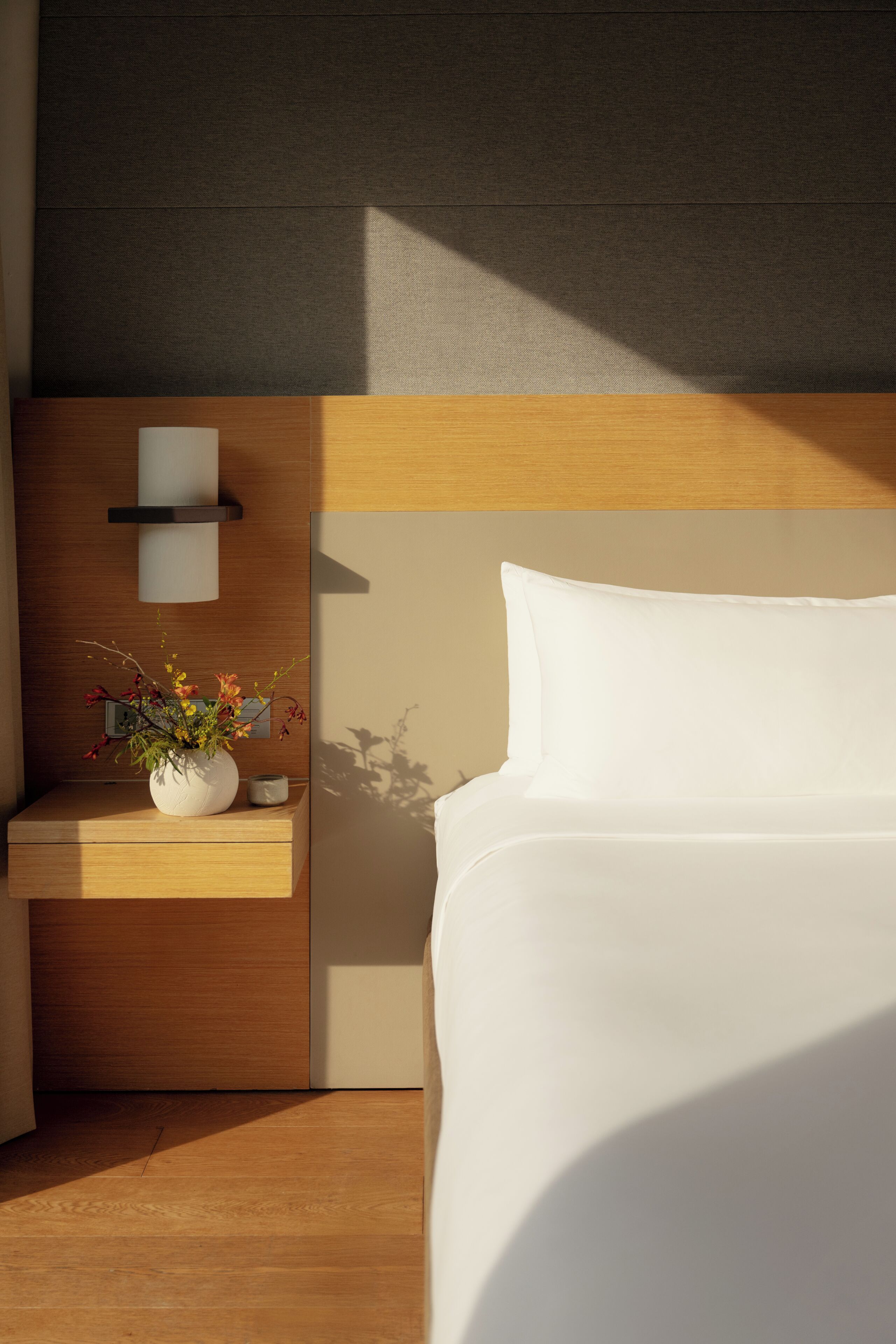 A modern hotel room with a wooden headboard, a bedside table with a vase of flowers, and a white bed.