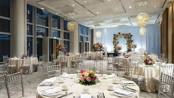 Un salón de banquetes con mesas redondas dispuestas para un evento formal, con centros de mesa florales y un gran arco floral al fondo.