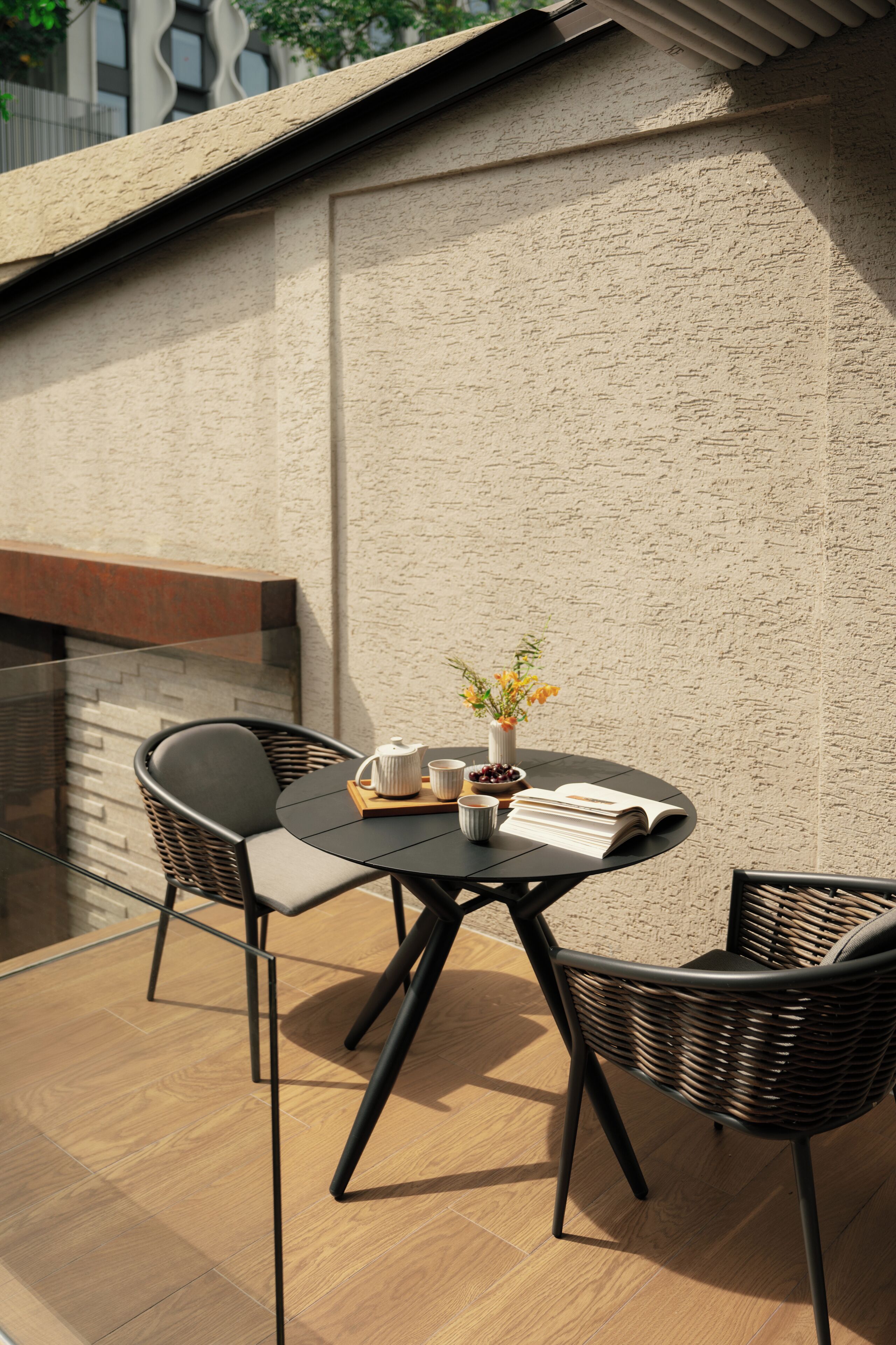 A round table with two chairs, a book, a teapot, and a vase with flowers on an outdoor patio.