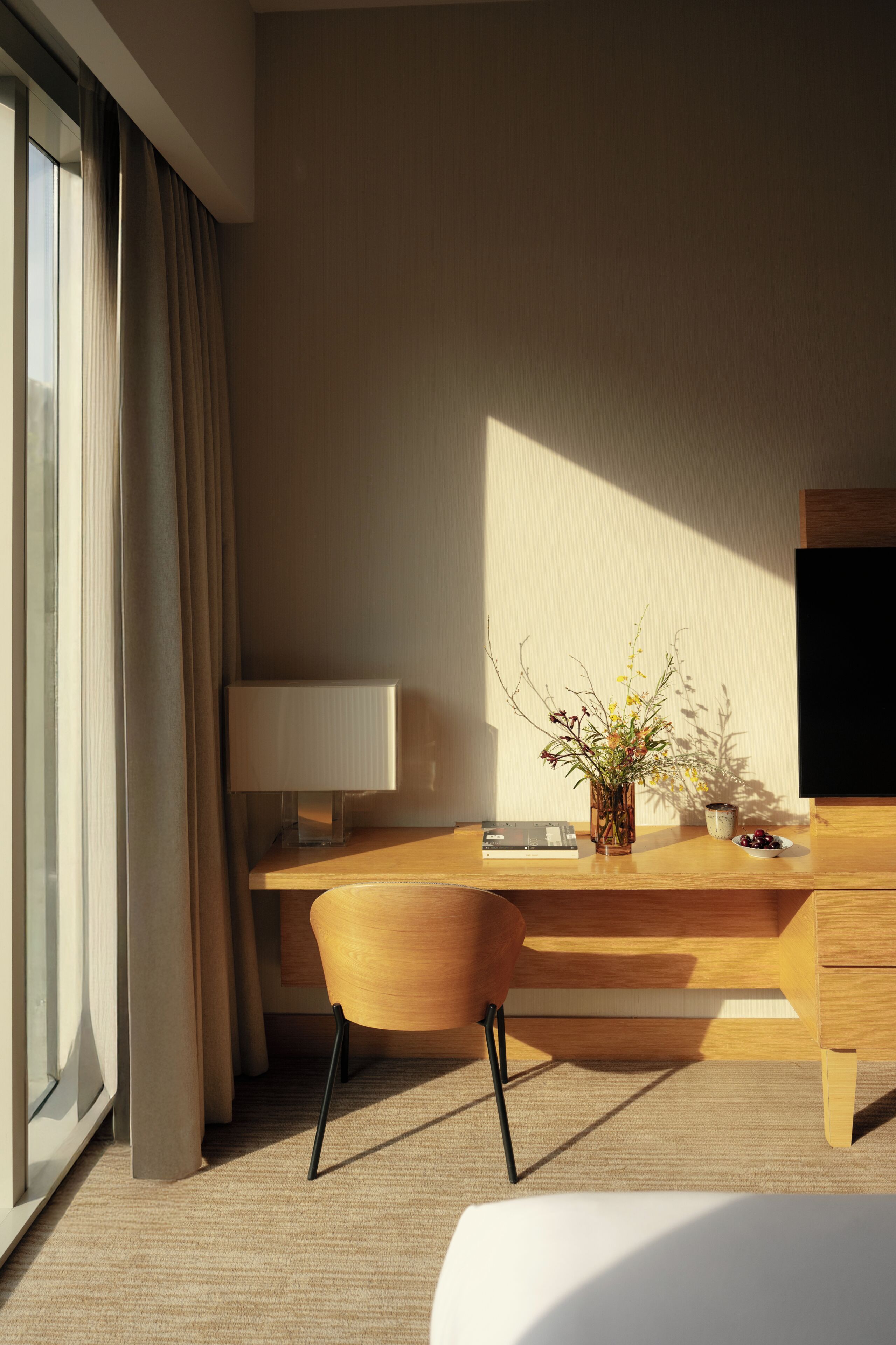 A modern hotel room with a wooden desk, a lamp, a vase with flowers, and a television.