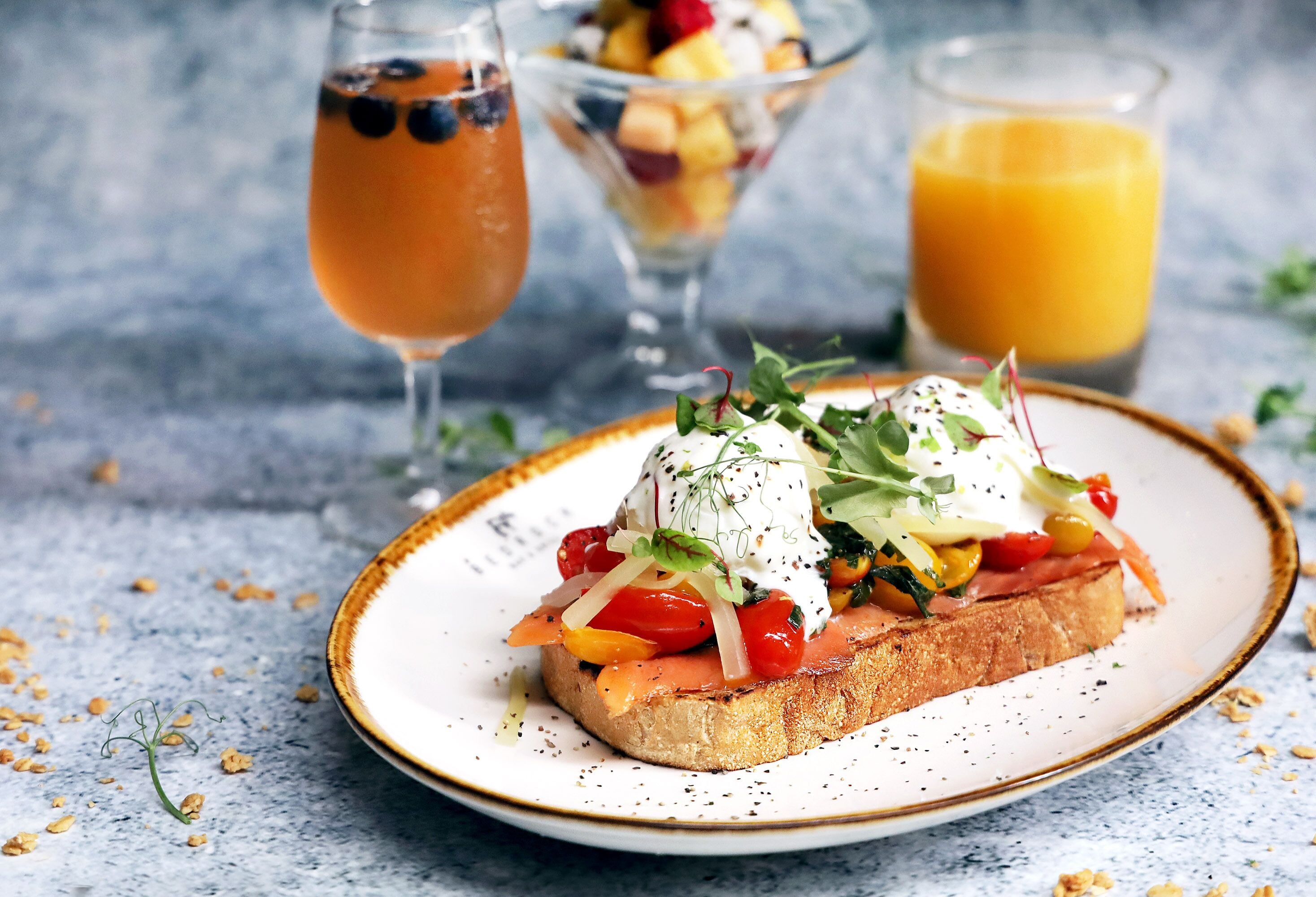 A plate with a sandwich topped with vegetables and a poached egg, accompanied by a glass of orange juice and a fruit bowl in the background.
