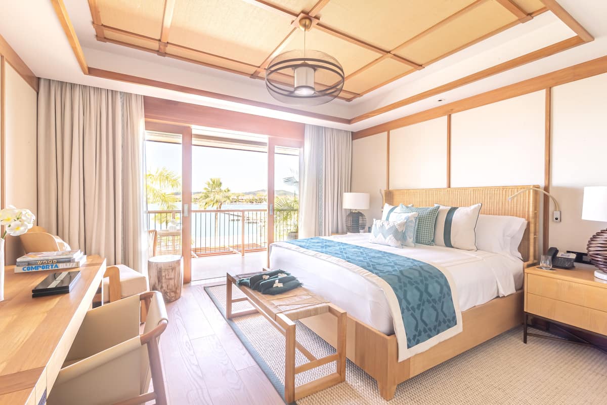 A hotel room with a large bed, a desk with a chair, a bedside table, and a view of palm trees outside.