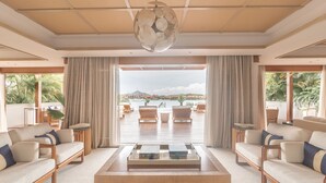 A spacious living room with a large window offering a view of a beach and boats. There are two sofas, a coffee table, and a hanging light fixture. - L'Escale Resort Marina & Spa (Mahé Island)