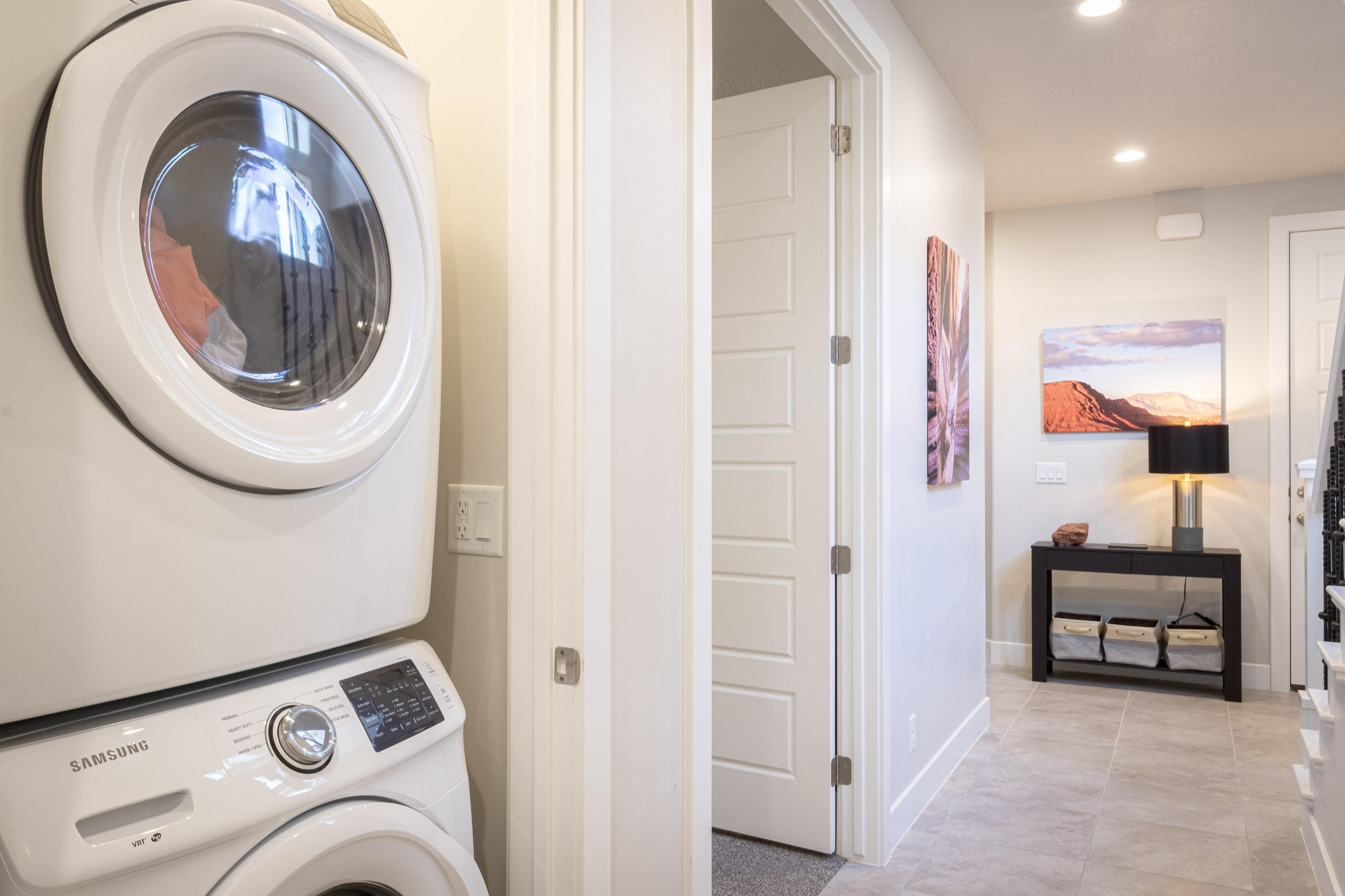 A laundry room with a Samsung washer and dryer, a door leading to a hallway with a painting and a lamp on a console table.