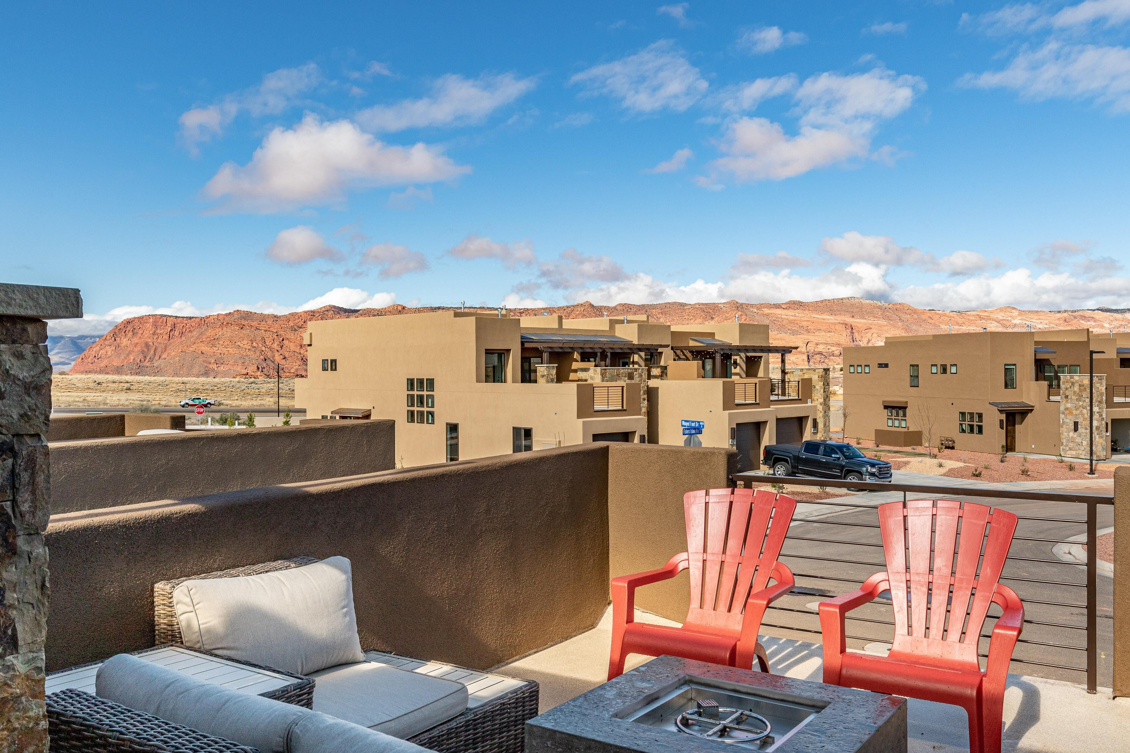 Une terrasse sur le toit, aménagée avec des meubles en osier et des fauteuils Adirondack rouges, offrant une vue sur un paysage désertique avec des bâtiments en arrière-plan.