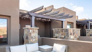 An outdoor patio with wicker furniture and stone pillars, under a pergola with string lights. - Solara Escape (St. George)