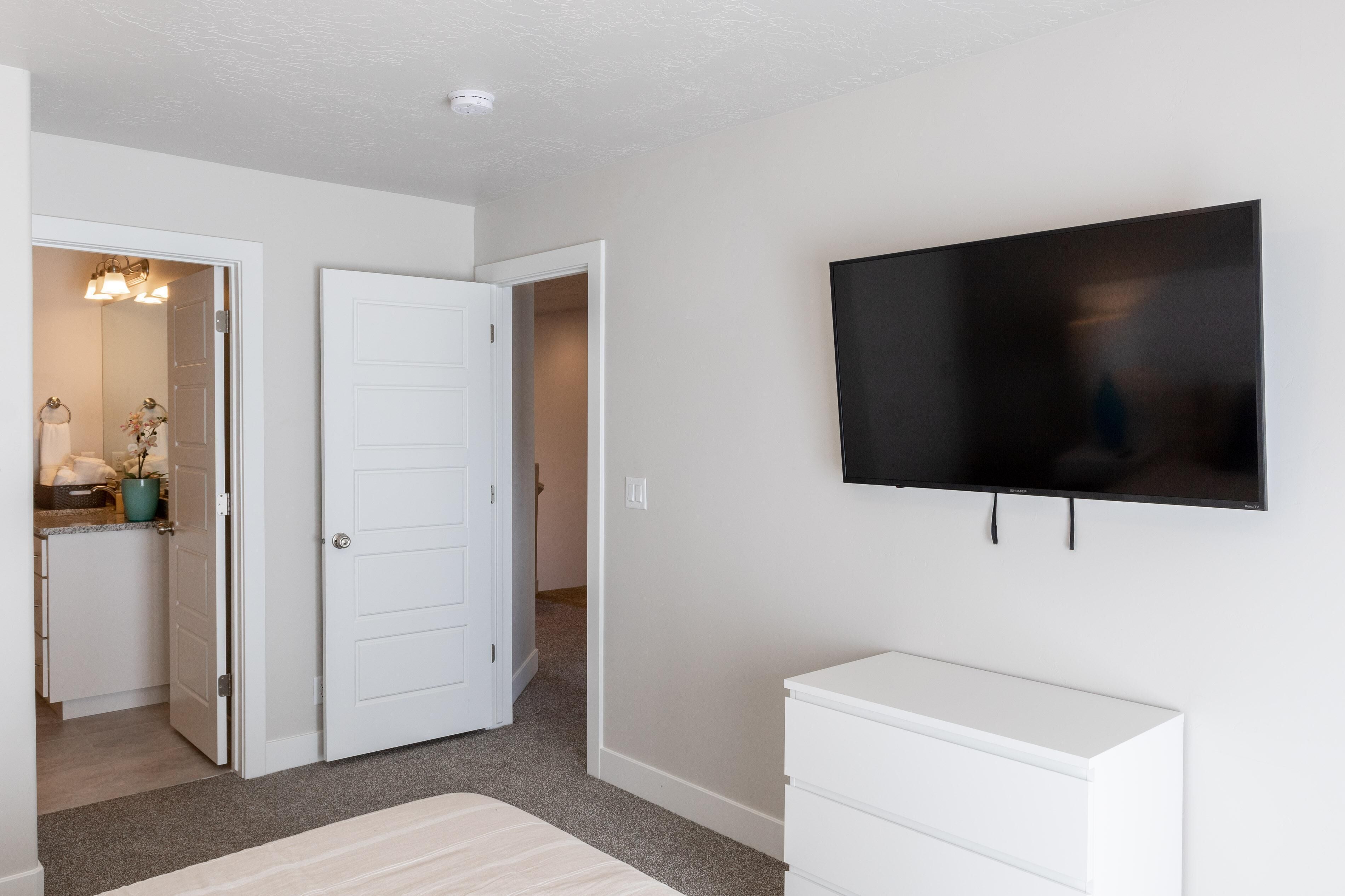 A room with a white dresser, a flat-screen TV mounted on the wall, and a door leading to a bathroom with a sink and mirror.