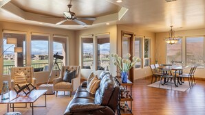 A spacious living room with a leather sofa, a coffee table, and a dining area with a table and chairs. Large windows offer a view of the mountains. - Chip Shot (St. George)