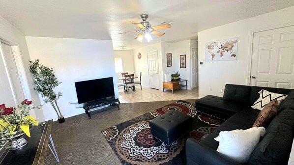 A living room with a flat-screen TV, a black leather sofa, a patterned rug, and a ceiling fan.