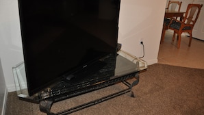 Un televisor de pantalla plana sobre un pedestal de vidrio en una habitación con piso alfombrado y sillas de madera al fondo.