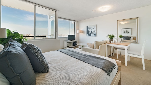 Ein modernes Schlafzimmer mit einem großen Bett, einem Schreibtisch und einem Sessel. Es gibt einen Fernseher, eine Pflanze und ein Fenster mit Blick.
