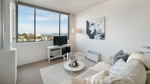 Ein modernes Wohnzimmer mit großem Fenster, einem Flachbildfernseher, einem Glastisch, einem weißen Sofa und Blick auf die Stadt.