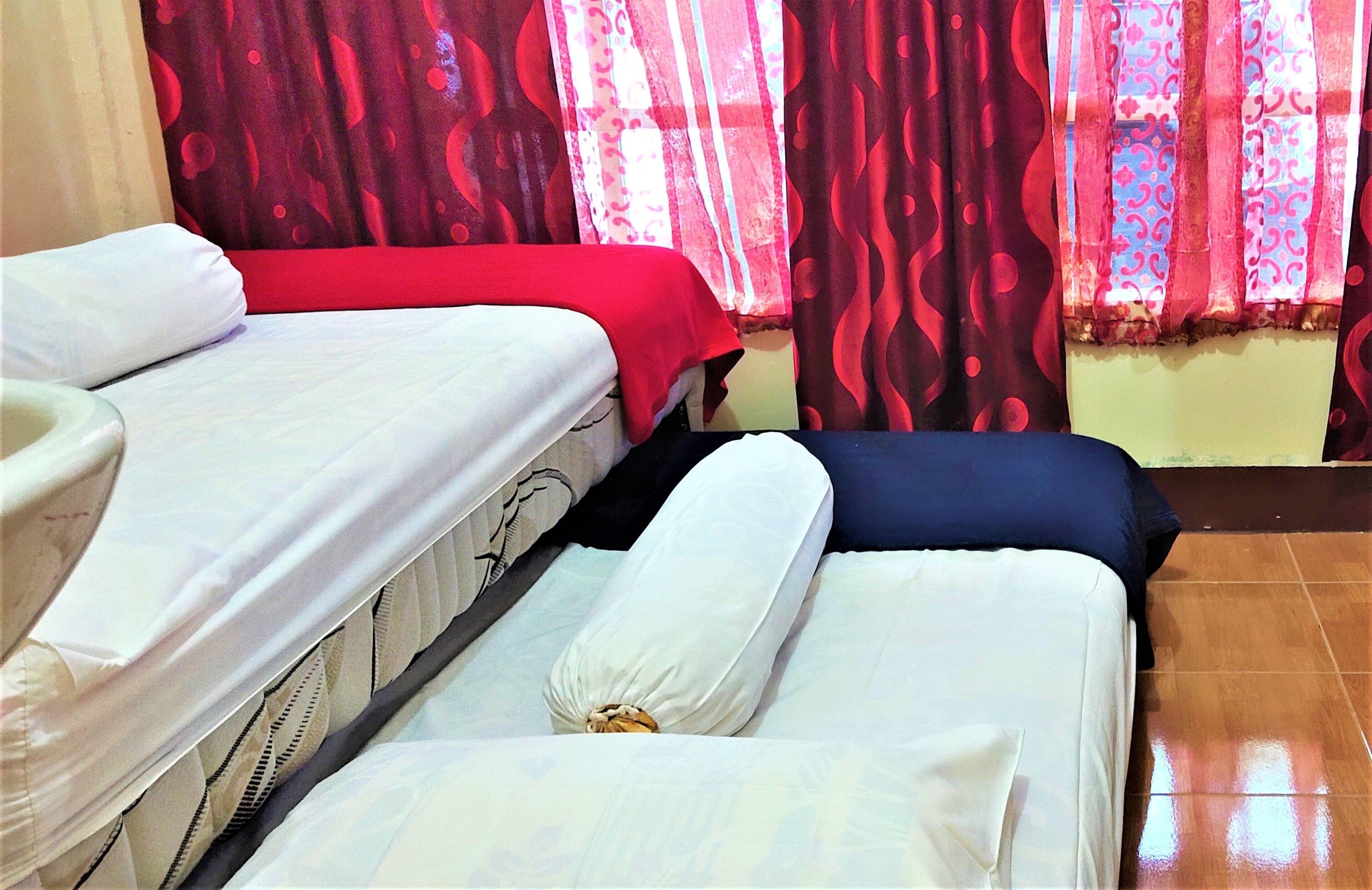 A hotel room with two beds, each with white bedding and a red and blue bedspread. The room has a tiled floor and red curtains with a pattern.