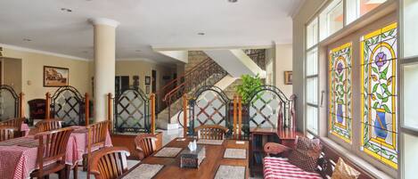 A dining area with a wooden table, chairs, and a bench. There is a staircase with a railing and a view of the outside through large windows.