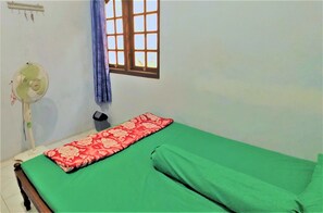 A single bed with green bedding, a red patterned blanket, a standing fan, a window with blue curtains, and a wall-mounted light fixture. - Wisma Artha (Ngaglik)