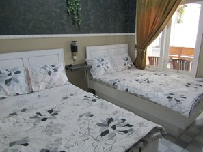 Two beds with floral bedding, a bedside table with a lamp, and a view of the outdoors through a window with curtains. - Penginapan Melati Sarangan (Plaosan)