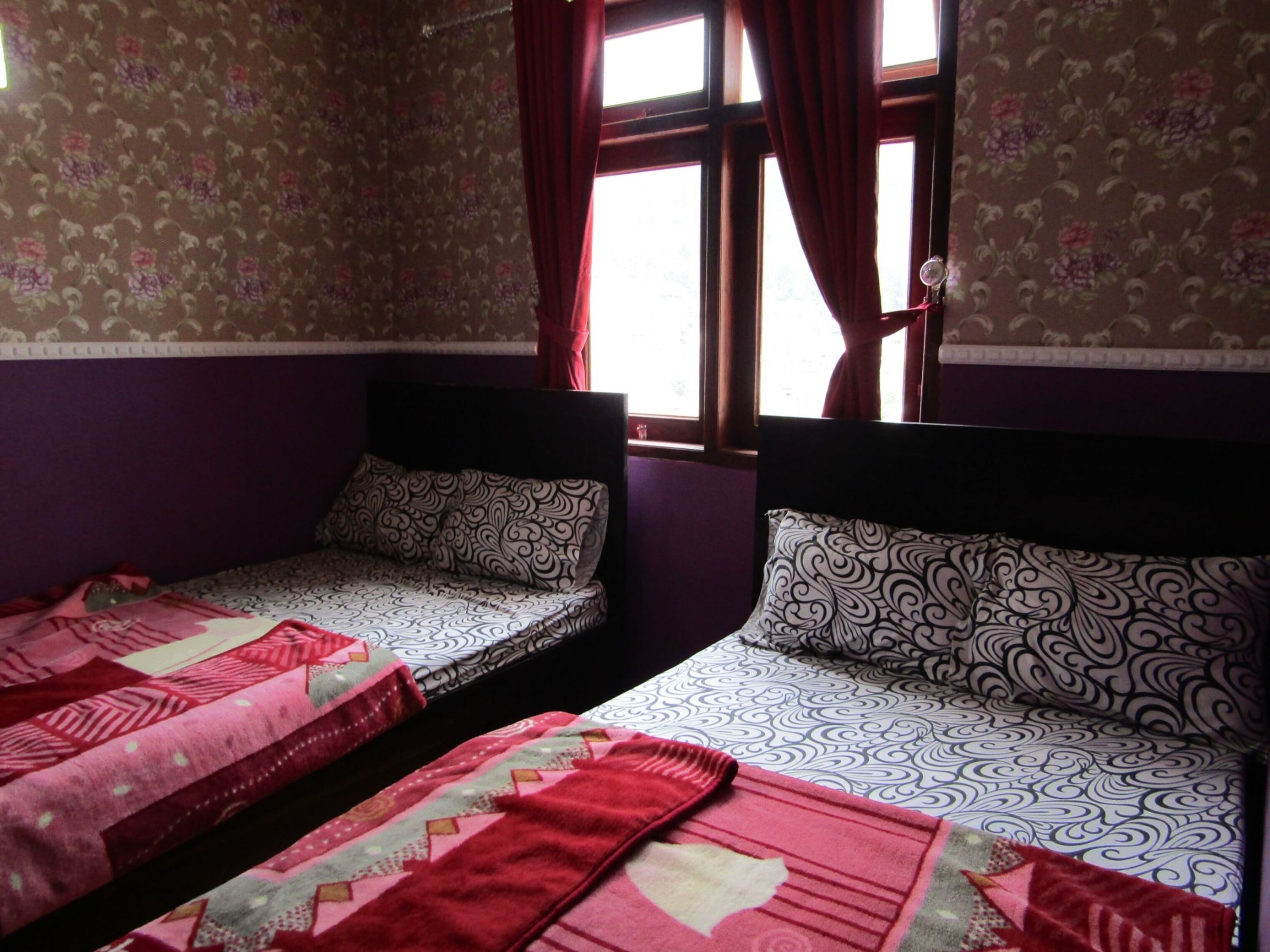 A room with two beds, each with patterned bedding and pillows. There is a window with curtains, and the walls have a floral wallpaper pattern.