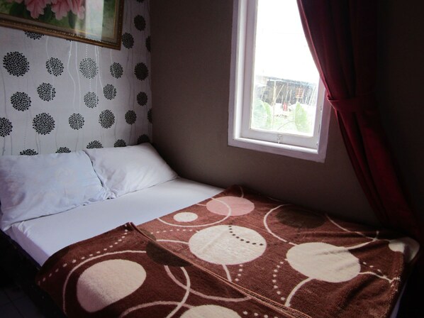 A single bed with a white pillow and a brown blanket with circular patterns, a window with a view of greenery, and a curtain partially drawn.