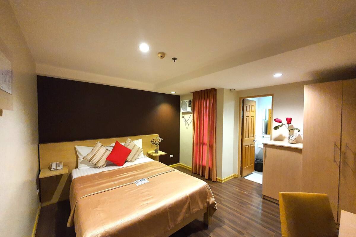 Quarto de hotel com cama, mesa de cabeceira, escrivaninha, cadeira e guarda-roupa.