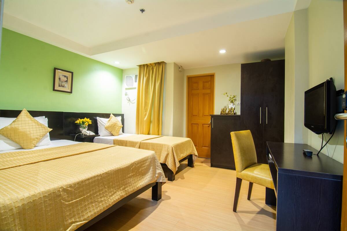 Quarto de hotel com duas camas, uma escrivaninha, uma cadeira, uma televisão e um guarda-roupa.