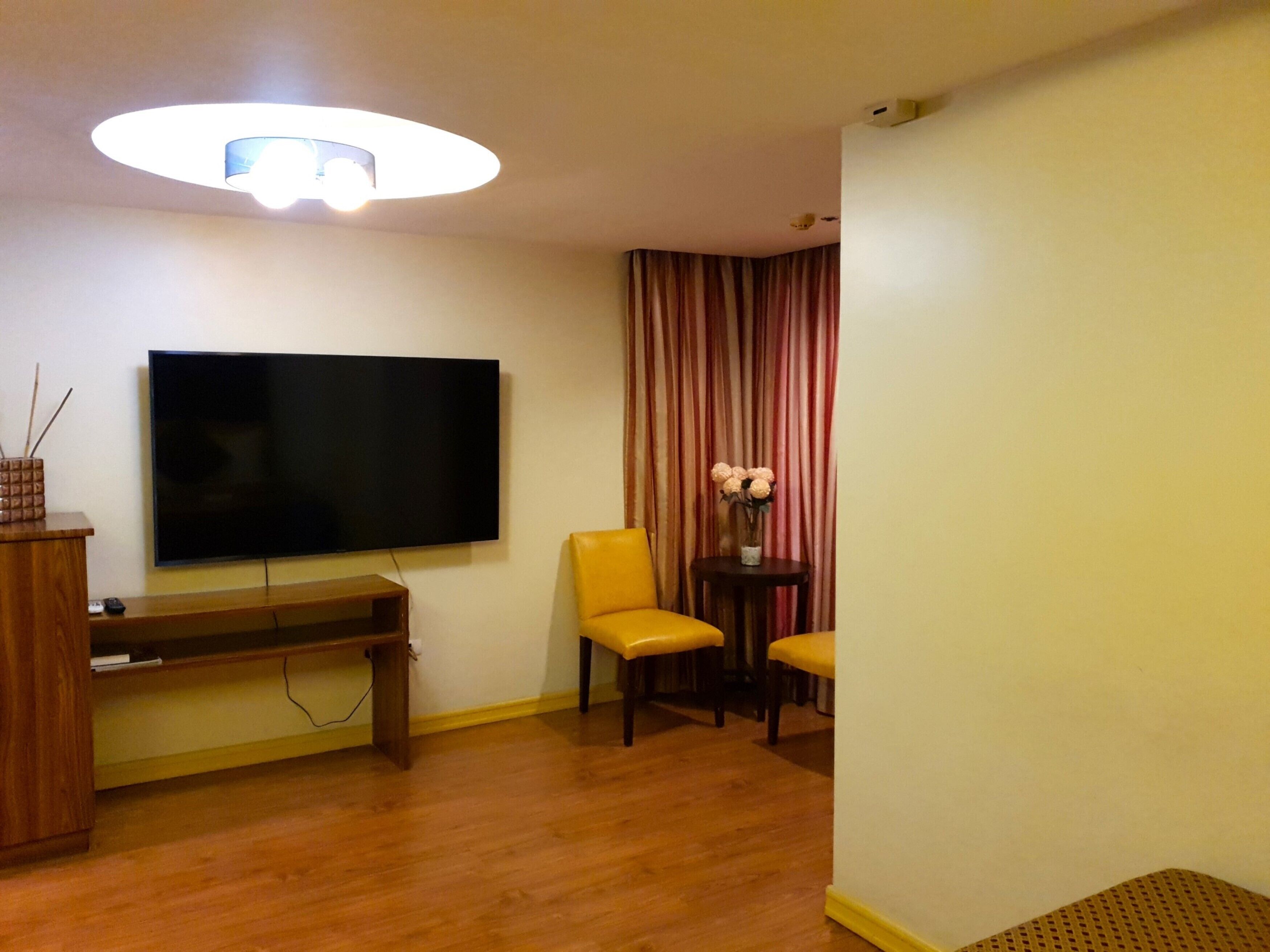 A hotel room with a flat-screen TV mounted on the wall, a wooden bench, two yellow chairs, a small table with a vase of flowers, and a ceiling light fixture.