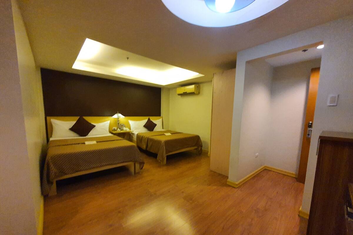 Quarto de hotel com duas camas, um claraboia, piso de madeira e uma porta que dá para outro quarto.