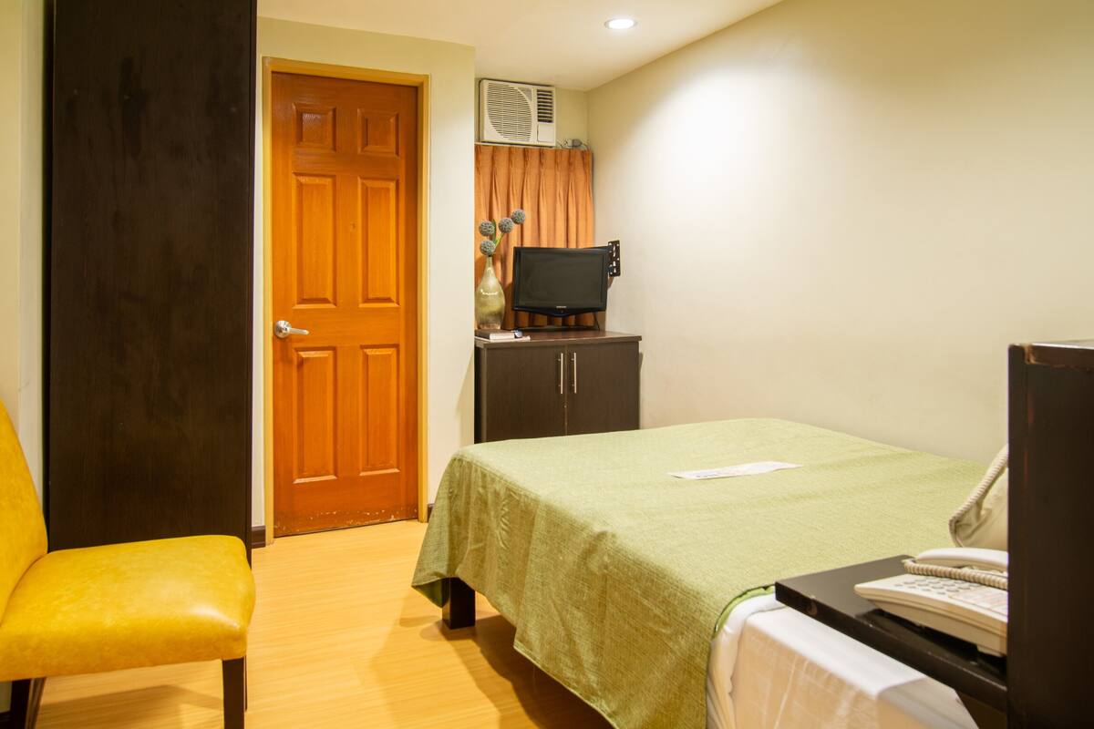 Quarto de hotel com uma cama, uma mesa com telefone, uma cadeira, uma televisão sobre um móvel e um aparelho de ar condicionado.