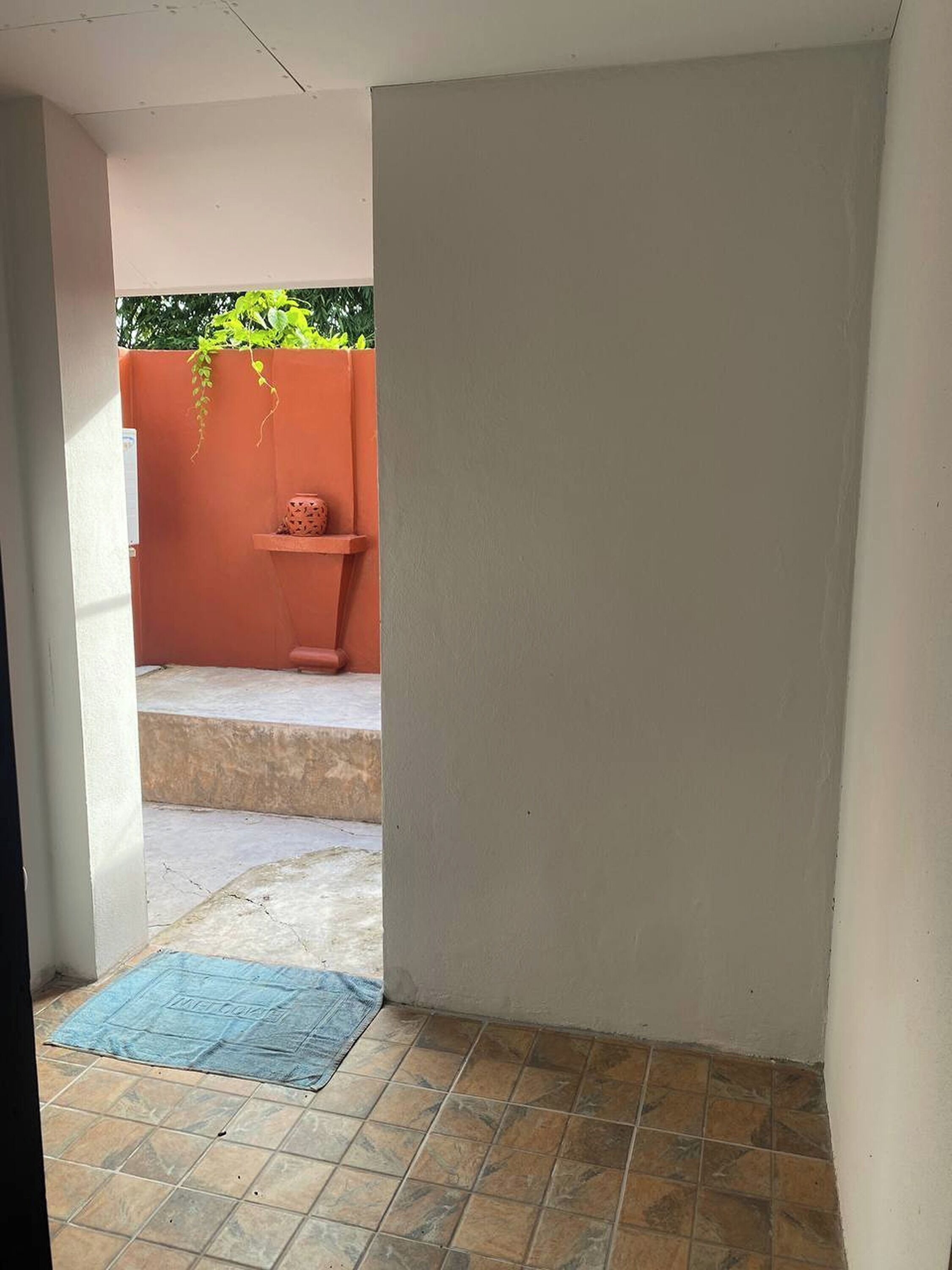Een smalle, open corridor met betegelde vloer, een kleine trede die leidt naar een verhoogd terras, en een bloempot op het terras.