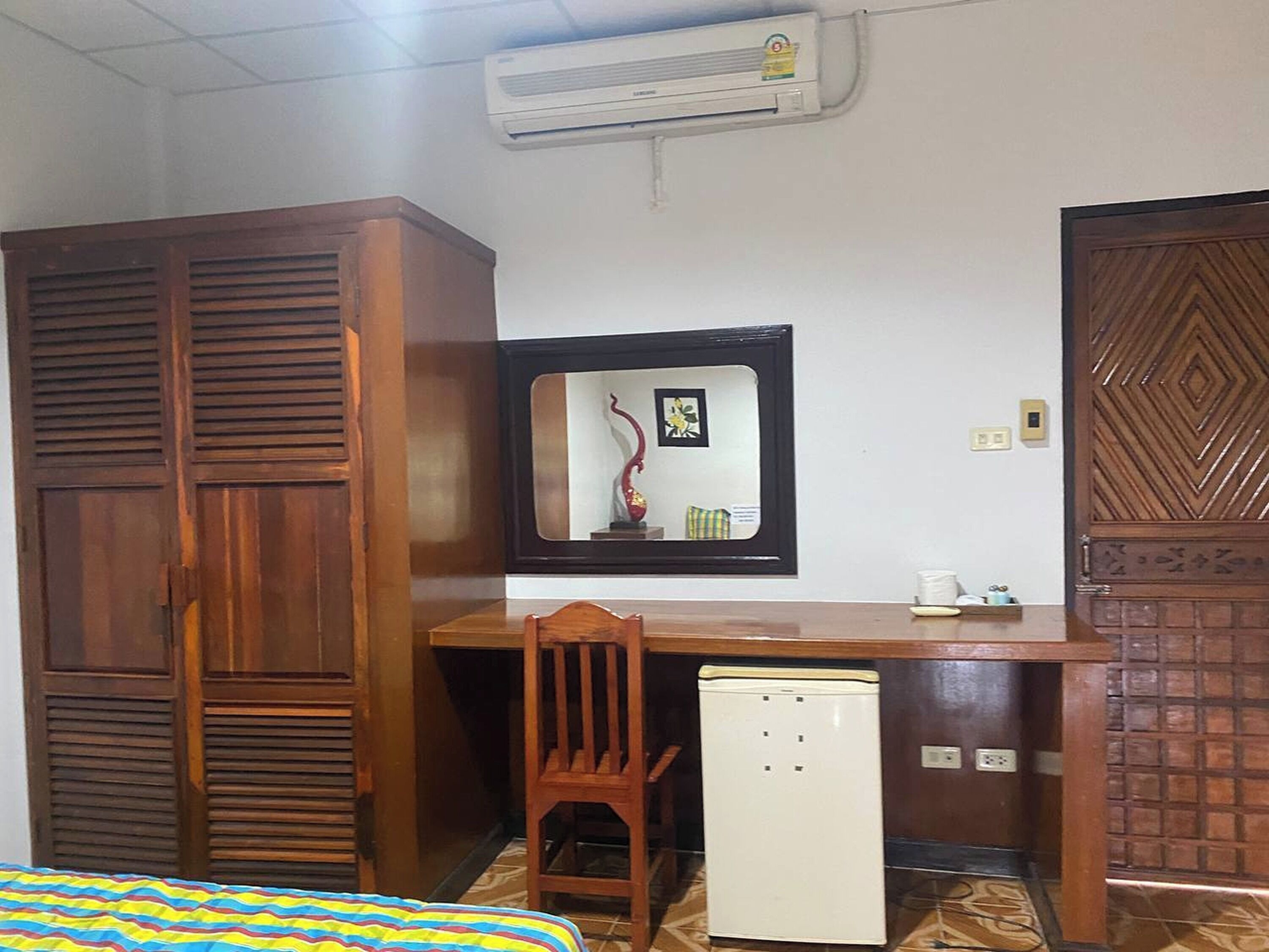 A room with a wooden wardrobe, a desk with a mirror, a small refrigerator, and a bed with a striped bedspread.