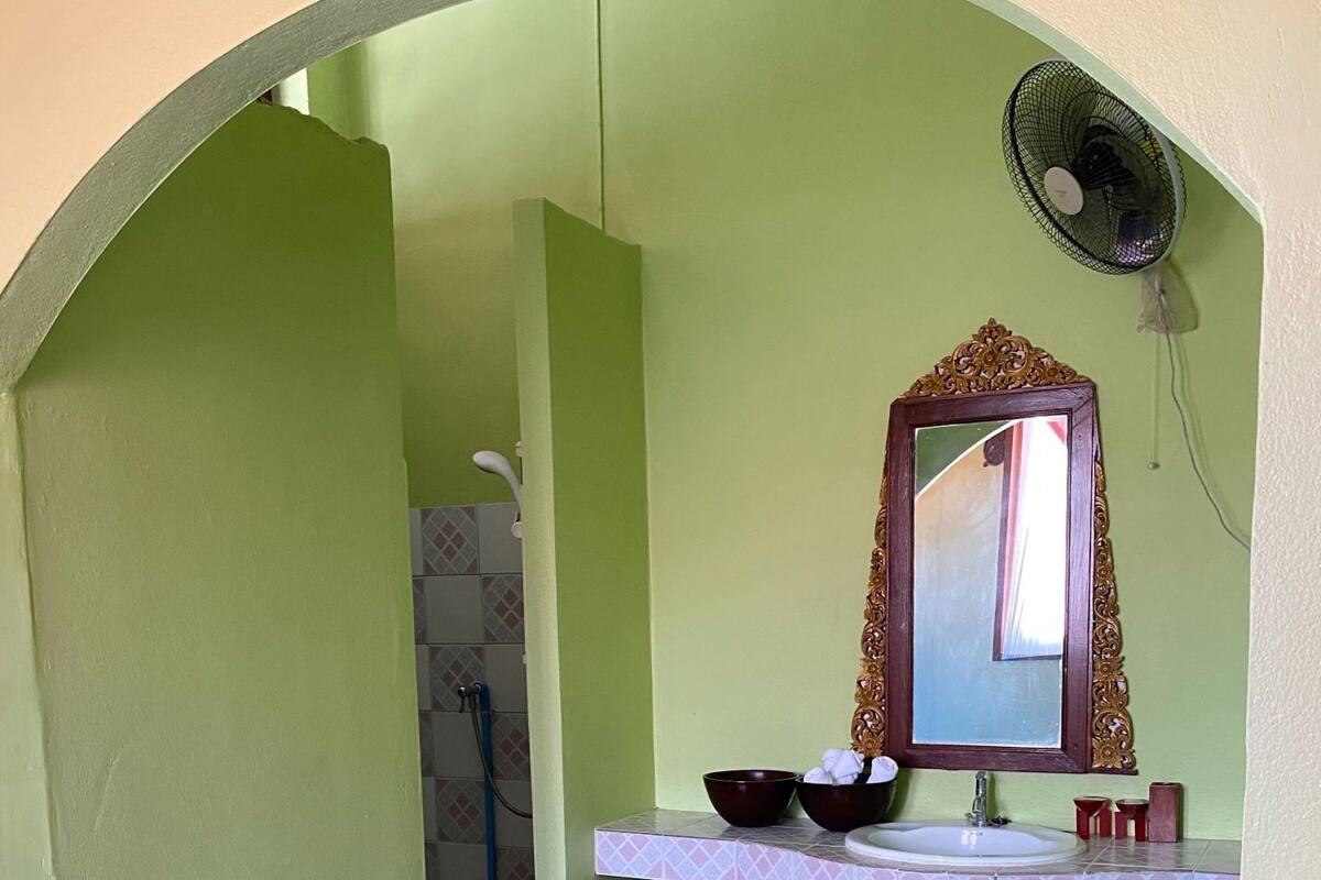 A bathroom with a green wall, a wooden vanity, a mirror, a sink, a fan, and a shower area.