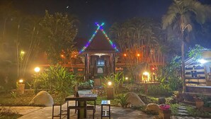 Um gazebo com luzes coloridas, cercado por plantas tropicais e palmeiras.