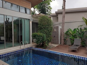 A modern outdoor pool area with a glass door entrance, a small seating area, and various plants. - Areeca Mahogany Zone (Choeng Thale)