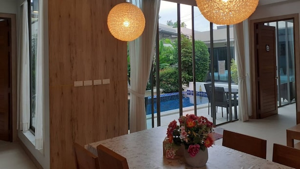 A modern dining area with a large table, wooden accents, and large windows offering a view of a pool and greenery. - Areeca Mahogany Zone (Choeng Thale)