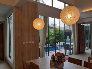 A modern dining area with a large table, wooden accents, and large windows offering a view of a pool and greenery. - Areeca Mahogany Zone (Choeng Thale)