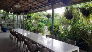 A covered outdoor dining area with a long white table and wicker chairs, surrounded by lush greenery and hanging plants. - Areeca Mahogany Zone (Choeng Thale)