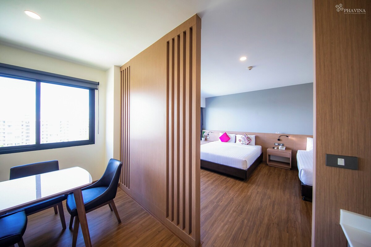 A hotel room with a wooden floor, a bed with white bedding and a pink pillow, a bedside table with a lamp, and a window with blinds.