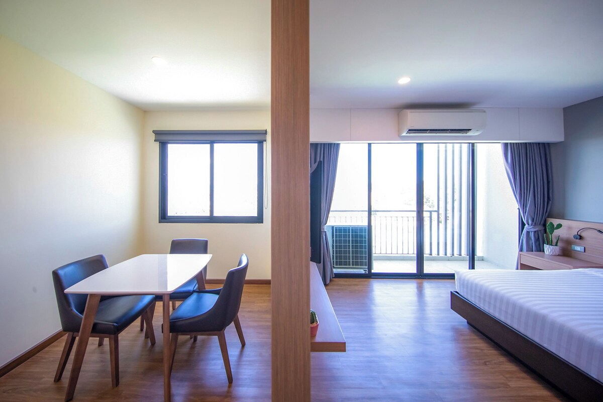 Quarto de hotel com piso de madeira, uma cama, uma escrivaninha com cadeiras, uma janela com cortinas e um aparelho de ar condicionado.