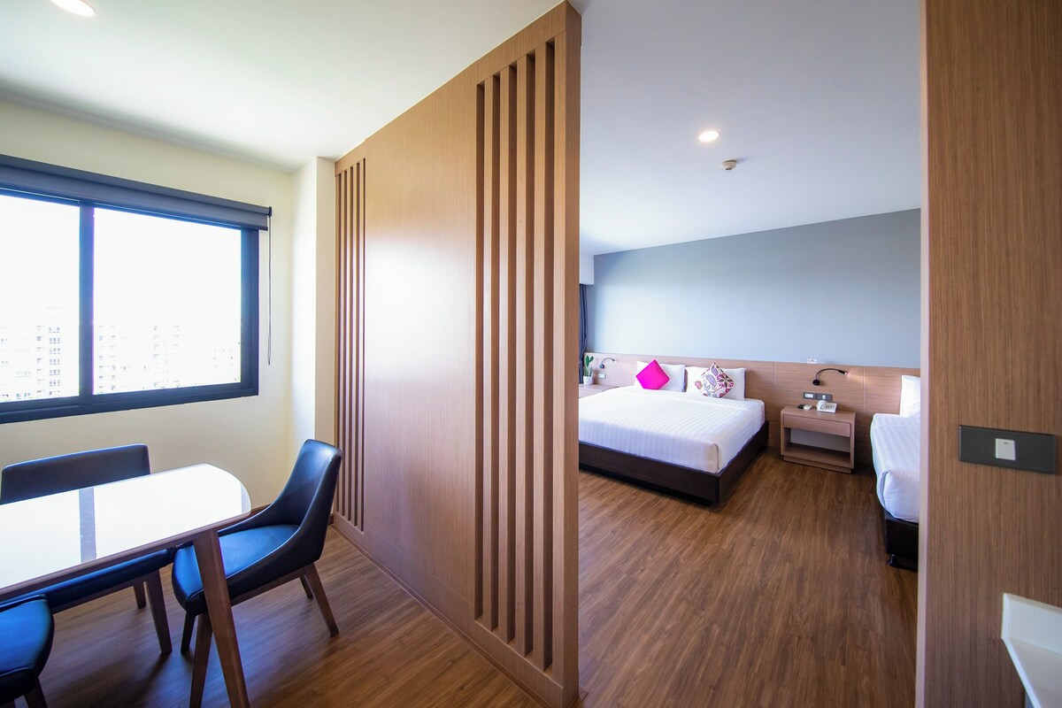 A hotel room with a wooden floor, a bed with white bedding and a pink pillow, a bedside table with a lamp, and a window with blinds.