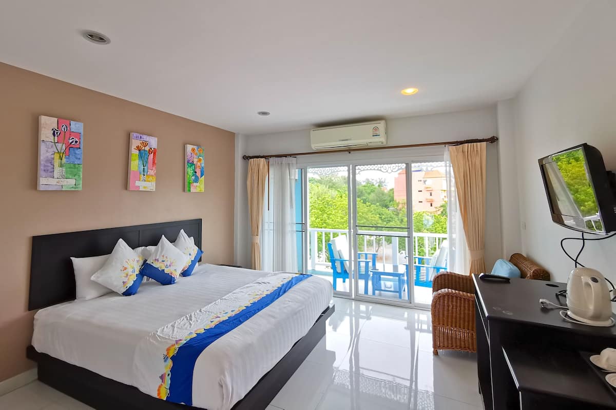 A hotel room with a bed, a TV, a kettle, and a balcony with blue chairs.