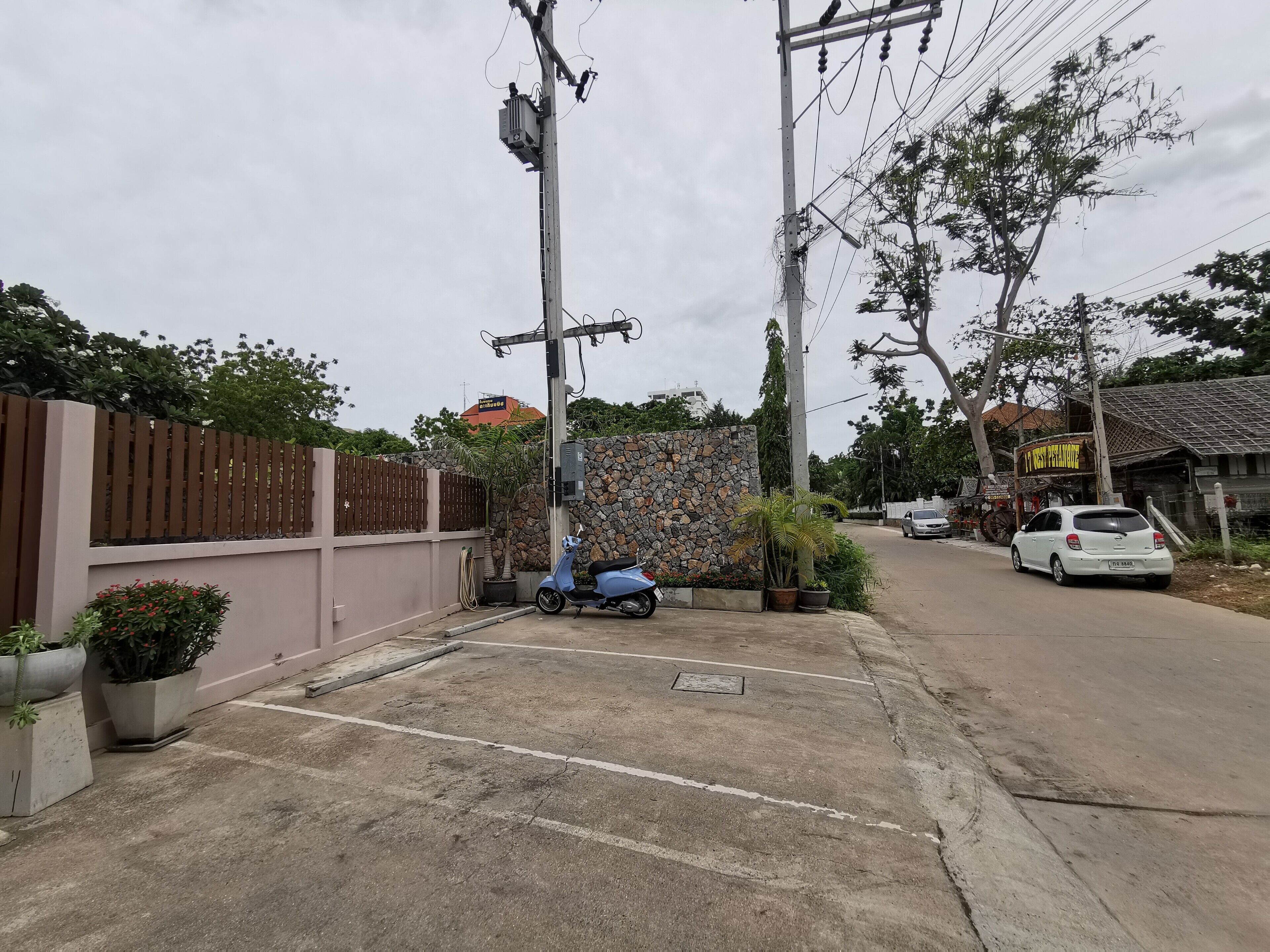 Un carrer residencial amb una moto aparcada, un cotxe blanc i una paret de pedra amb una tanca metàl·lica.